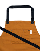 The Folded Steel Chef Apron features eco-friendly rPET canvas in brown, a large front pocket with a white label, black adjustable neck strap, and diagonal waist tie—an essential sustainable kitchen accessory shown on a white background.