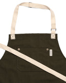 The Folded Steel Chef Apron with Eco-Friendly rPET Canvas Construction is dark green with beige straps, a large front pocket featuring two buttons, and a small right-side tag—all shown on a white background.
