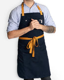 A person with visible forearm tattoos wears a light blue shirt and the Folded Steel Chef Apron, made from eco-friendly rPET canvas. The dark apron features tan straps, waistband, and a chest pocket; hands are clasped at chest level.