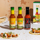 Three bottles from Elijah's Xtreme Mild & Hot Trio Hot Sauce Bundle are displayed on a table next to soft chicken tacos with onions and cilantro. The Elijah's Xtreme product box and tortillas appear in the background. Enjoy mild and hot sauces with every bite.