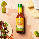 A bottle from the Elijah’s Xtreme Mild & Hot Trio Hot Sauce Bundle sits on a light textured surface, next to a taco with cilantro and onions, lime wedges, and corn tortillas.