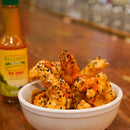 A white bowl of saucy chicken wings with black and white sesame seeds sits on a wooden table. In the background is a bottle from Elijah’s Xtreme Mild & Hot Trio Hot Sauce Bundle by Elijah’s Xtreme.