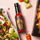 A bottle of Elijah's Xtreme Regret Reserve Hot Sauce by Elijah's Xtreme sits on a pink surface beside lime wedges, an orange pepper, and nachos topped with beef, cheese, cilantro, and sour cream.