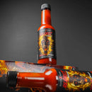 A bottle of Elijah's Xtreme Regret Reserve Hot Sauce by Elijah's Xtreme with its fiery skull label stands upright next to its box against a dark background, while another Carolina Reaper hot sauce bottle is partially visible in the foreground.