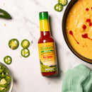 A bottle of Elijah’s Xtreme Roasted Cayenne and Jalapeño Pepper Sauce by Elijah's Xtreme rests on white marble beside sliced jalapeños and a bowl of creamy soup topped with swirling hot sauce, with a green cloth peeking into the corner.