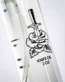 A close-up of a clear Folded Steel Knife Oil bottle from Folded Steel—2 oz size, ideal for outdoor cooking and high carbon blades—with a dagger piercing a rose among leaves on the label, plus black measurement lines along the side.