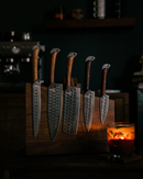 The Folded Steel Lotus 5-Piece Knife Set AUS10 Stainless Steel, with wooden handles, is displayed on a wooden knife block. A lit candle in a glass holder nearby casts a warm glow in the dimly lit room.