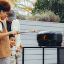 A woman uses a pizza peel to slide a pizza into a Gozney Arc Lite Outdoor Pizza Oven while a man and a child watch.