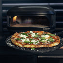 A fresh pizza topped with melted cheese and basil sits on a black peel in front of a Gozney Arc Lite Outdoor Pizza Oven with a visible flame.