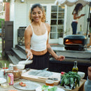 A woman smiles while preparing authentic pizza at an outdoor table next to a Gozney Arc Lite Outdoor Pizza Oven.