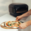 A person prepares a pizza with sauce, cheese, and basil on a wooden peel in front of a Gozney Arc Lite Outdoor Pizza Oven.