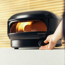 A hand adjusts the dial on a black Gozney Arc Lite Outdoor Pizza Oven, with a flame visible inside its dome.