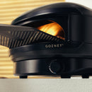 A black Gozney Arc Lite Outdoor Pizza Oven with a visible flame in the cooking chamber and a metal pizza peel inserted.