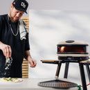 A person sprinkles flour onto a 16-inch pizza near a Gozney Arc XL Outdoor Pizza Oven with a visible flame inside.