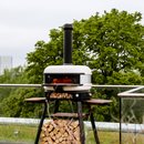 White outdoor pizza oven with a Gozney Dome Flue Extension for improved airflow, sitting on a black stand filled with stacked firewood on a balcony.