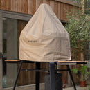 The Gozney Dome Gen 2 Cover by Gozney is a beige, water-repellent cover that protects a triangular outdoor pizza oven, shown on a black metal stand near a wooden building and potted plant.
