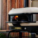 The Gozney Dome Gen 2 Mantel, a modern outdoor pizza oven by Gozney with a digital display, visible flame, and sleek stainless steel shelf, sits on a black stand against a wooden wall.