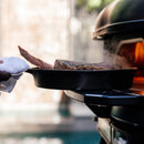A hand uses a cloth to place a cast iron skillet with steak onto the Gozney Dome Gen 2 Mantel by Gozney, steam rising from the meat as the skillet rests partly on the stainless steel shelf of the outdoor pizza oven.