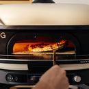 A hand uses a pizza peel to remove a golden, leopard-spotted pizza with melted cheese from the Gozney Dome Gen 2 Neapolitan Arch by Gozney, which delivers excellent heat retention for perfect baking.