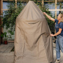 A person pulls a water-repellent, beige Gozney Dome Gen 2 Series Stand Cover over a large outdoor oven, with plants and a modern building visible in the background.