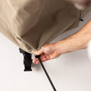 A person pulls a black cord through a grommet on the beige Gozney Dome Gen 2 Series Stand Cover, tightening the water-repellent fabric securely around a wheel or leg of an object on a white surface.