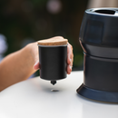 A hand placing a Gozney Dome Steam Injector with a cork lid into a circular opening on a black stove for artisan bread baking.