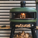Olive green Gozney Dome Wood pizza oven with kiln-dried hardwood burning inside and logs stored in the base.