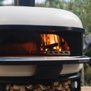 A white, dome-shaped Gozney Dome Wood outdoor pizza oven with kiln-dried hardwood burning inside the hearth and a stack of firewood stored in the base.