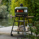 Olive green Gozney Dome Wood pizza oven with a fire burning inside, set on a metal stand with kiln-dried hardwood stored on the lower shelf.