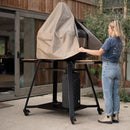 A woman in a blue t-shirt, jeans, and beige boots stands outside by a wooden building, revealing a Gozney Dome XL Gen 2 pizza oven covered with the water-repellent Gozney cover on a wheeled black metal stand.