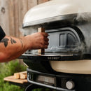 A tattooed person opens a steaming Gozney Dome XL Gen 2 Door outdoor oven, with wood pieces nearby and greenery behind—ideal for smoking and slow-roasting.