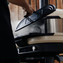Someone lifts the insulated Gozney Dome XL Gen 2 Door from a modern Gozney outdoor pizza oven, releasing steam, with a wooden wall in the background.