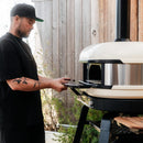 A man in a black shirt and cap uses the Gozney Dome XL Gen 2 Mantel pizza oven by Gozney near a wooden wall, creating an ideal pizza oven setup with plants in the background.