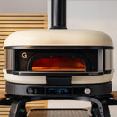 The Gozney Dome XL Gen 2 Neapolitan Arch, a modern cream pizza oven by Gozney, features high heat retention and a digital temperature display. It sits on a black stand with glowing interior and vent pipe rising above wooden slats in the background.