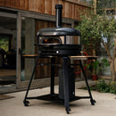 The Gozney Dome XL Gen 2 Neapolitan Arch, featuring high heat retention and a sleek black metal finish, stands on a wheeled cart with side shelves on a patio near plants, in front of a sliding glass door and wooden wall.