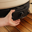 A person's hand is inserting or removing a black Gozney Wood-Fire Control Kit rechargeable battery pack with a large airflow control dial into a beige and black Gozney device on a wooden surface.