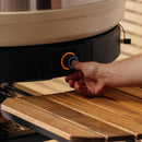 A hand adjusts the illuminated knob of the Gozney Wood-Fire Control Kit by Gozney, precisely managing airflow above a wooden slatted shelf.