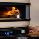 A close-up of the Gozney Wood-Fire Control Kit by Gozney shows precision airflow control, a digital display at 66°C, visible flames and wood inside the oven, and a hand reaching toward its side.