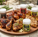 A wooden platter with grilled, sliced chicken, roasted lemon halves, herbs, garlic, and four jars of SALTOPIA Infused Sea Salts’ Grilled Chicken Bundle—Best Flavors for Chicken on the Grill—arranged outdoors on a sunlit table.