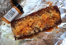 A rack of ribs coated with seasoning, brown sugar, and butter chunks rests on aluminum foil. Nearby on the foil is a bottle of Hardcore Carnivore Amplify Mega Savory Flavor Dust Seasoning from Hardcore Carnivore.