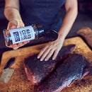 Someone sprinkles Hardcore Carnivore Black Beef Seasoning by Hardcore Carnivore onto a large raw beef cut on a wooden cutting board, preparing it for cooking. A knife lies nearby.