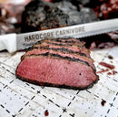 Several thick slices of smoked beef with a dark crust from Hardcore Carnivore Black Beef Seasoning and a pink interior are arranged in front of a large knife labeled Hardcore Carnivore on a white cutting surface with black markings.