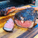 A large, grilled tomahawk steak rests on a cutting board, sliced to reveal its juicy medium-rare center. The rich crust and deep grill marks come from Hardcore Carnivore Black Beef Seasoning by Hardcore Carnivore. A grill sits in the background.