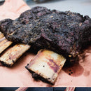 A slab of smoked beef ribs with a dark, seasoned crust from Hardcore Carnivore Black Beef Seasoning by Hardcore Carnivore rests on butcher paper, displaying exposed bones and tender, juicy meat.