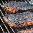 Close-up of two steaks with a charred crust on the grill, seasoned with Hardcore Carnivore Black Beef Seasoning by Hardcore Carnivore, bold grill marks visible on the metal grates.