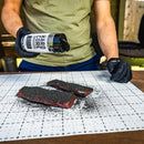 Wearing black gloves, a person sprinkles Hardcore Carnivore Black Beef Seasoning by Hardcore Carnivore—featuring activated charcoal—onto two raw meat slabs placed on a white grid cutting mat. Only their torso and hands are visible.