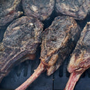 Several bone-in steaks with a dark, charred crust are grilling on the barbecue, seasoned with Hardcore Carnivore Black Beef Seasoning by Hardcore Carnivore. Smoke rises around the juicy steaks as they cook closely packed on the grill.