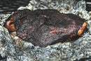 A close-up of a large, smoked beef brisket with dark, crispy bark rests on crumpled foil. The juicy meat is seasoned with Hardcore Carnivore Camo Game & Lamb Seasoning, highlighting visible smoke marks for mouthwatering appeal.