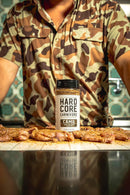 A person wearing a camouflage shirt stands behind a table with seasoned meat and a bottle of Hardcore Carnivore Camo Game & Lamb Seasoning by Hardcore Carnivore displayed prominently in the center.