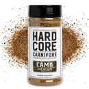 A container of Hardcore Carnivore Camo Game & Lamb Seasoning by Hardcore Carnivore is displayed with a burst of brown spice. The white label features bold black text and gold accents, making it an ideal BBQ rub for wild game and lamb.